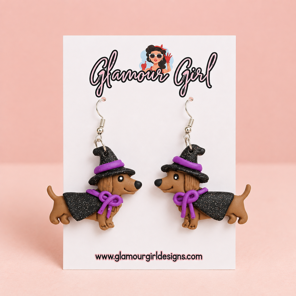 Purple witch dachshund dangle earrings with black hats and capes, purple bows and silver hooks on a white Glamour Girl card against a pale blush background.