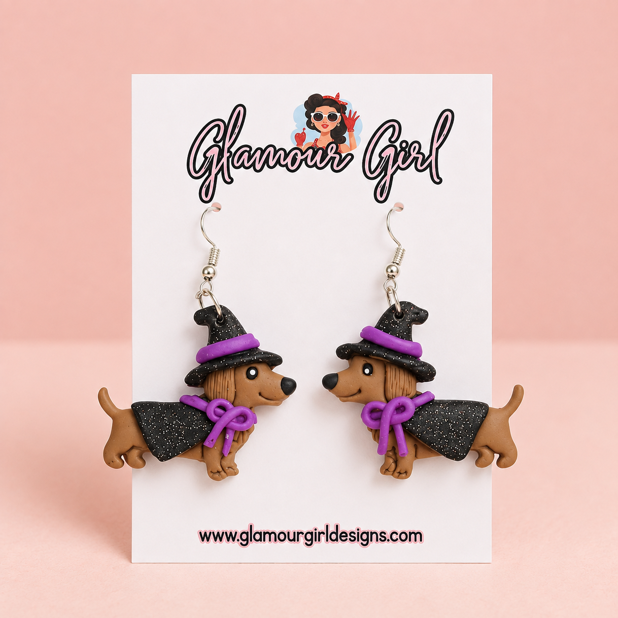 Purple witch dachshund dangle earrings with black hats and capes, purple bows and silver hooks on a white Glamour Girl card against a pale blush background.